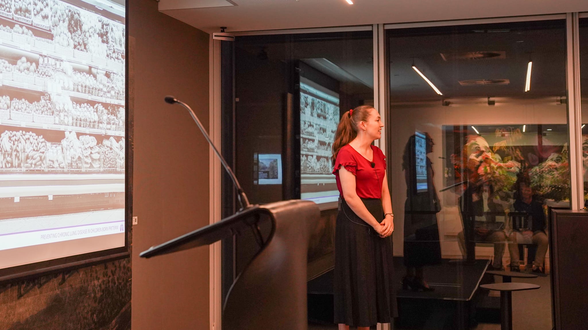 A woman standing in front of a large projector screen showing a supermarket produce section.