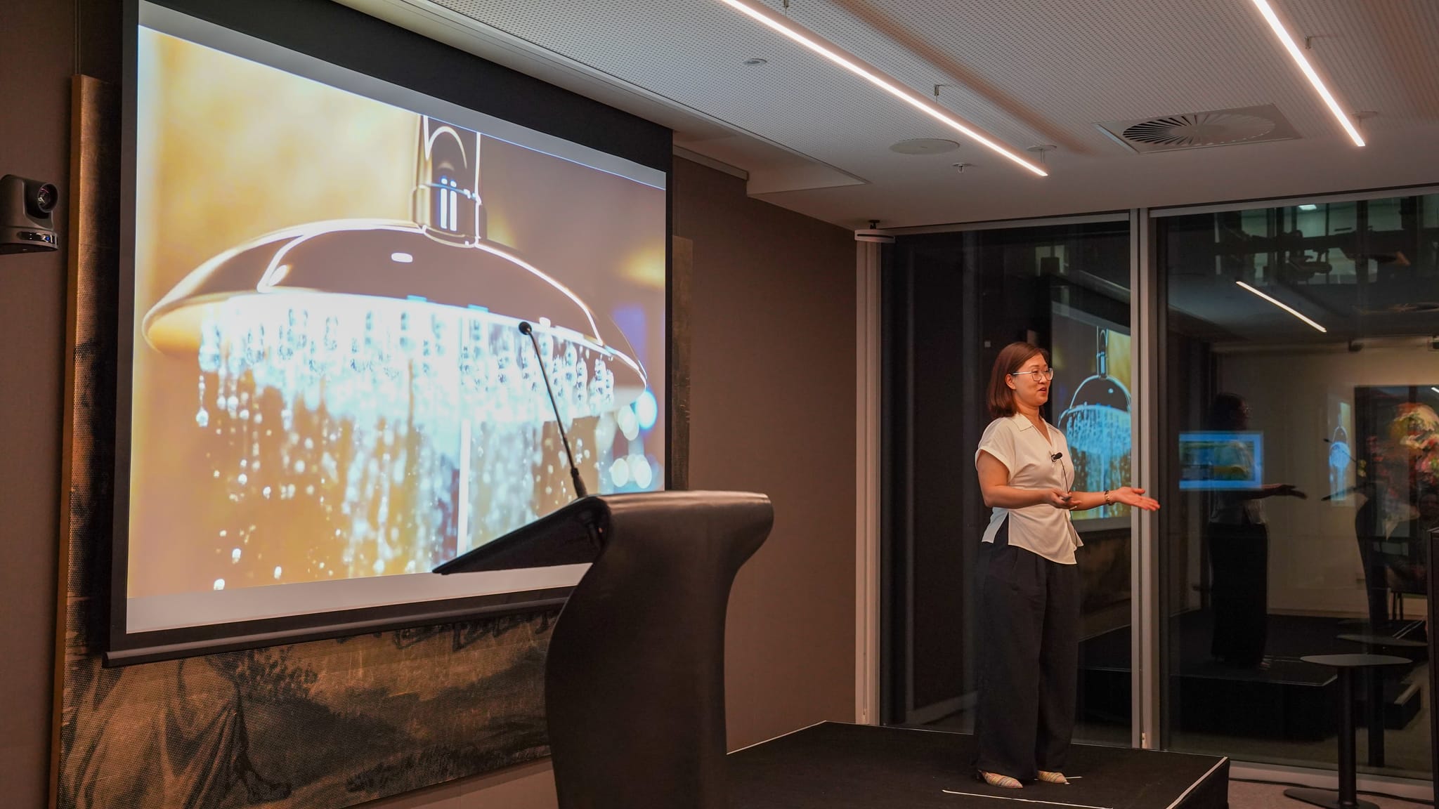 A female presenter in front of a large projector screen with an image of a shower head.